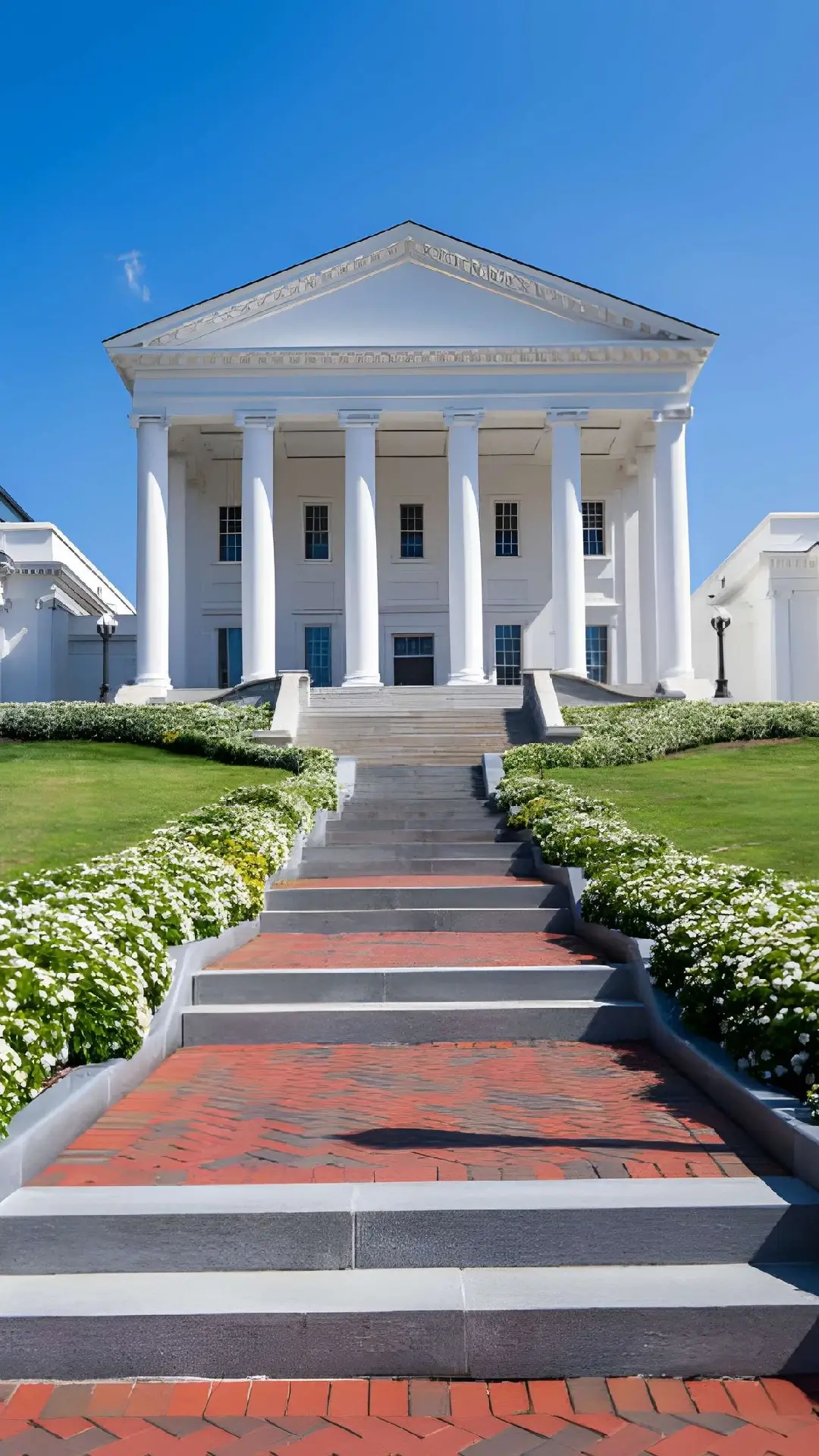 White neoclassical government building with tall columns, brick steps, and landscaped grounds under a clear blue sky.