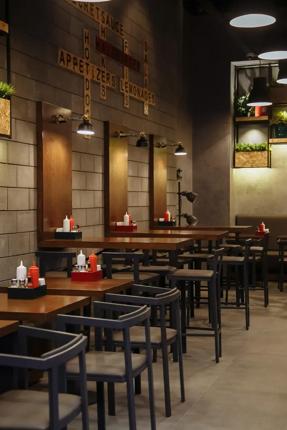 Modern restaurant interior with wooden tables, black chairs, pendant lighting, wall decor, and condiment stations.