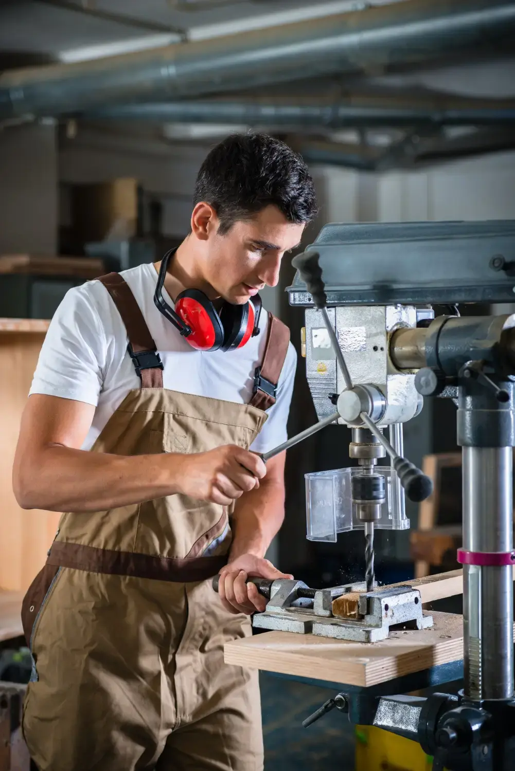 Skilled worker operating a drill press on a piece of wood in a woodworking workshop while wearing protective equipment.