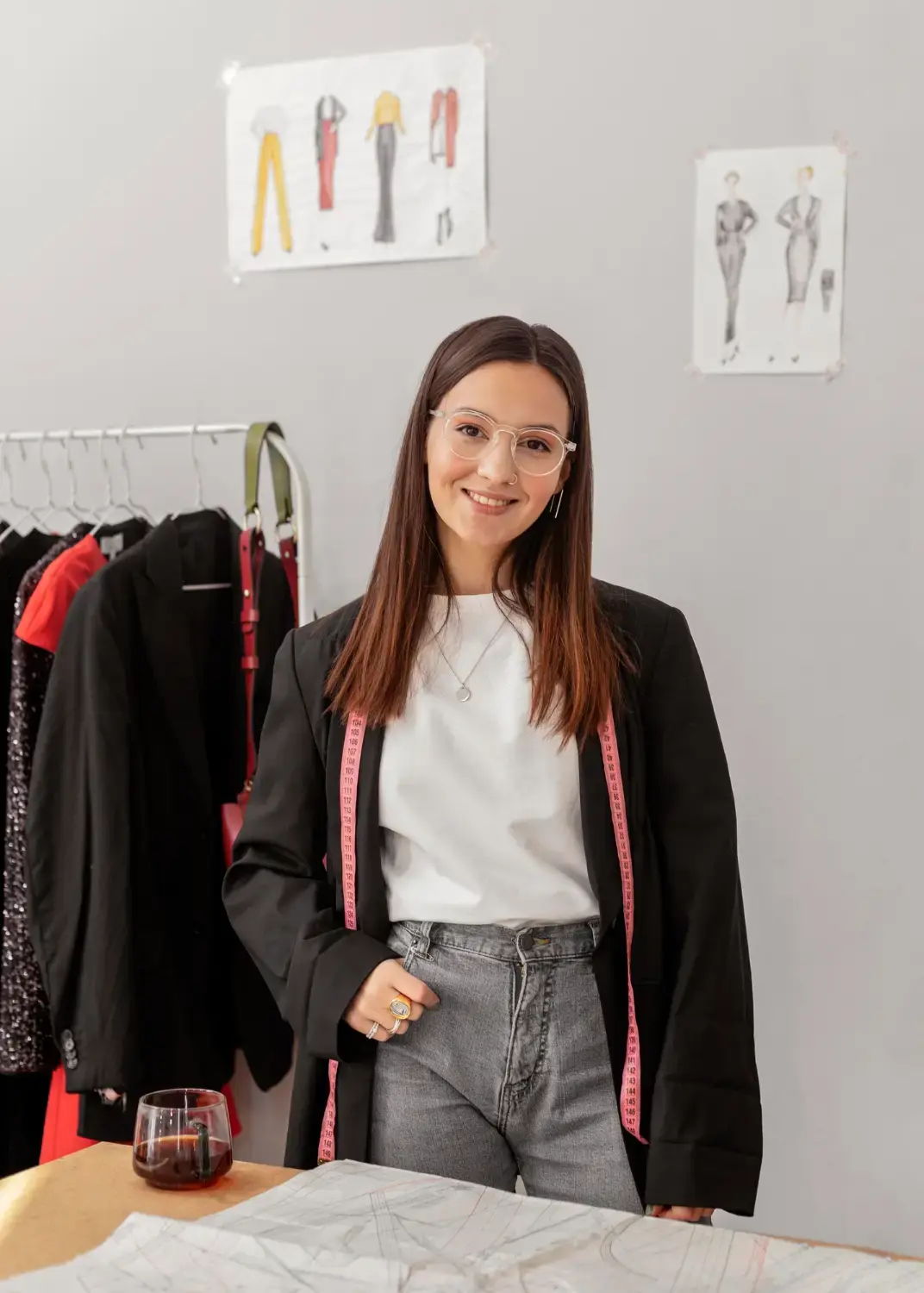 Female fashion designer standing in a clothing studio with a measuring tape, garment rack, fashion sketches, and sewing patterns.