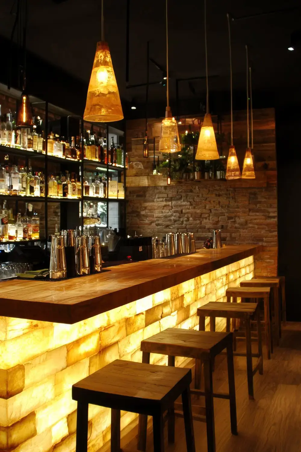 Modern bar interior with illuminated stone counter, wooden stools, pendant lights, and shelves of liquor bottles.