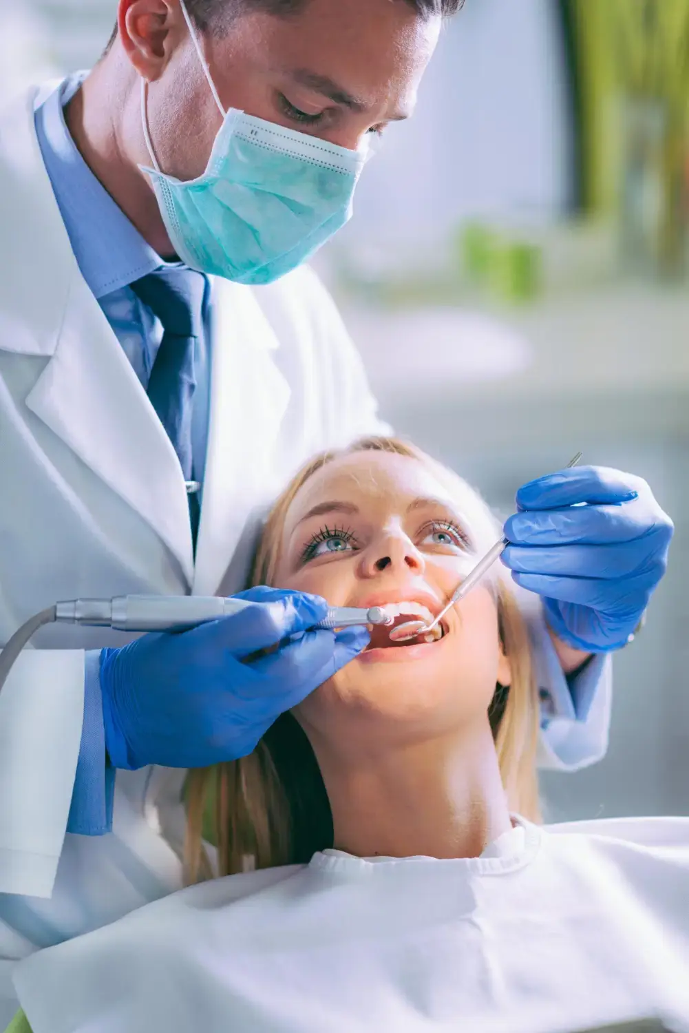 Dentist wearing a face mask and gloves examining a female patient’s teeth with dental instruments in a clinic.