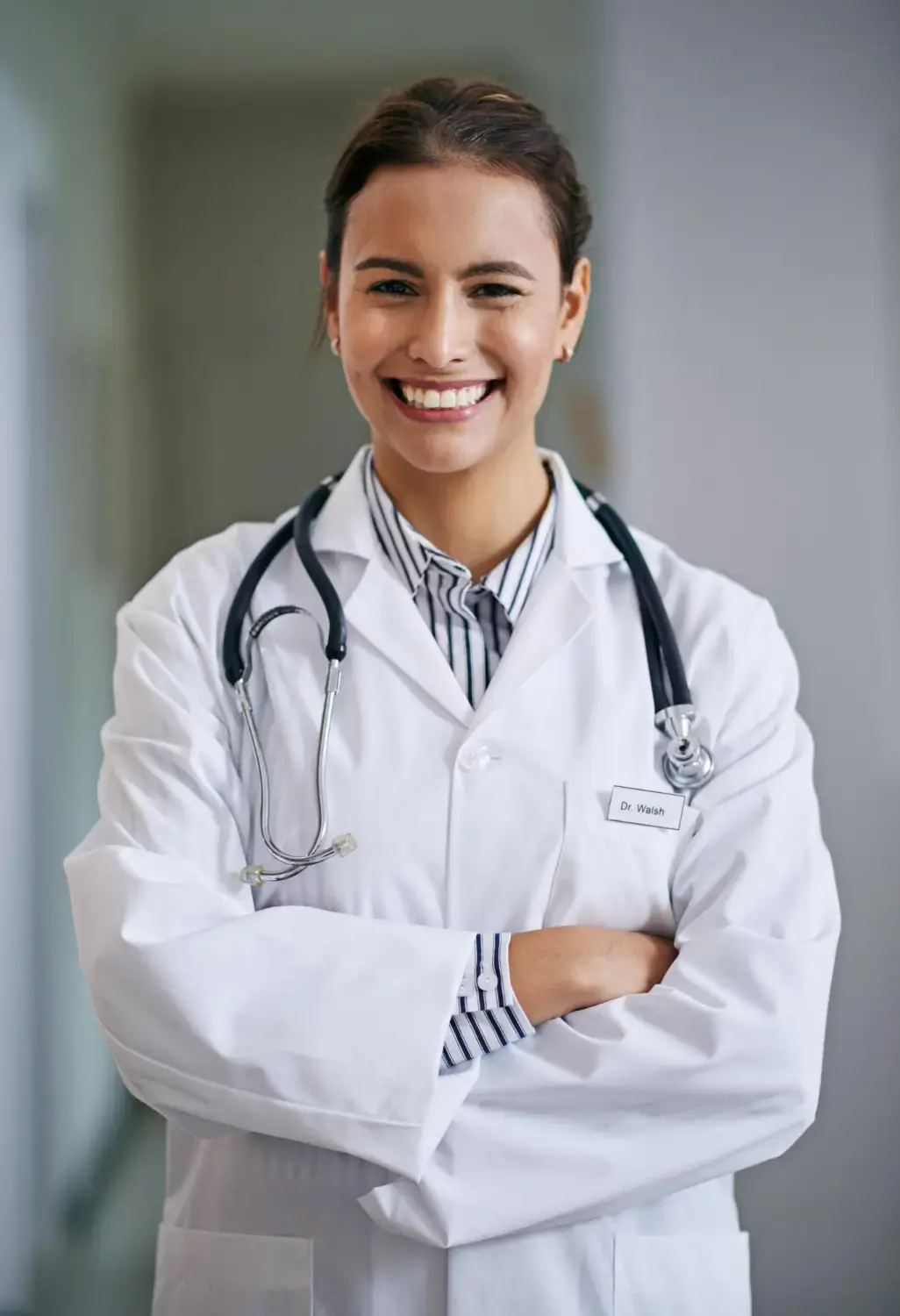 Smiling female doctor wearing a white coat and stethoscope standing with arms crossed in a medical clinic.