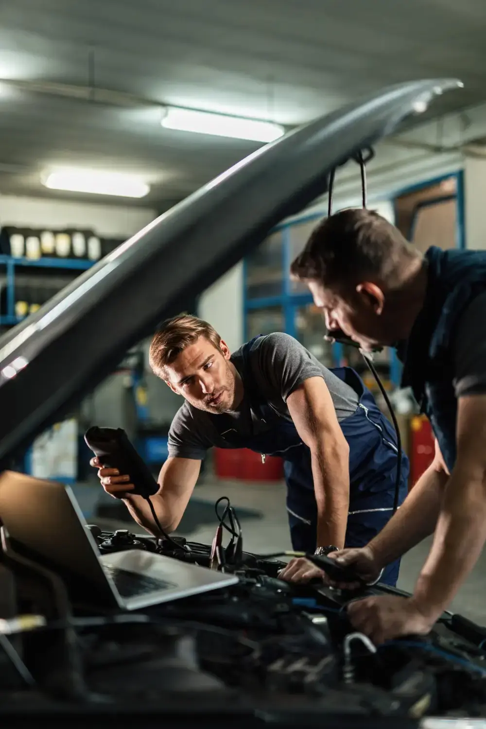 Two auto mechanics using diagnostic equipment and a laptop to inspect a car engine in an automotive repair shop.