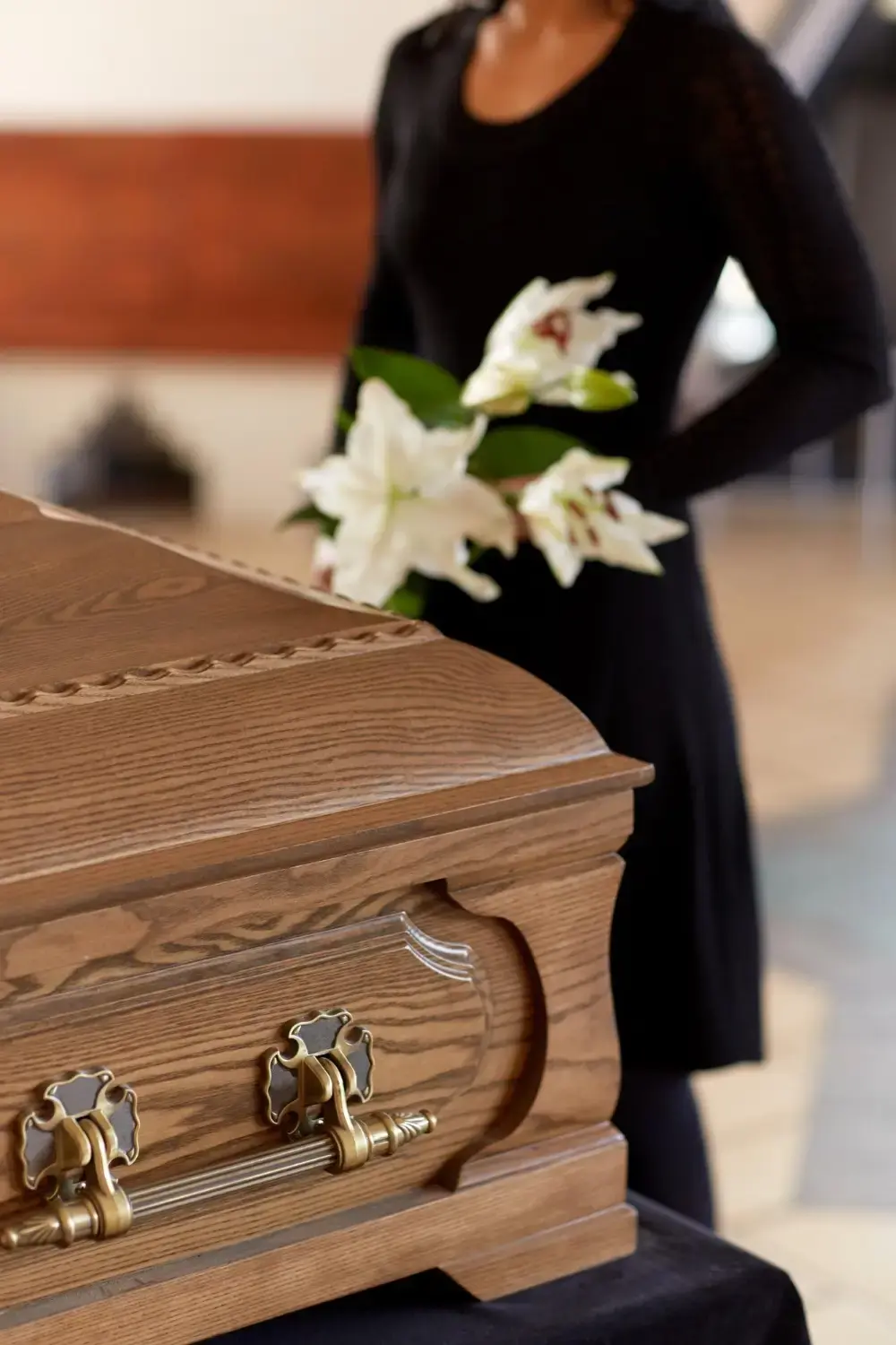 Wooden casket with white lilies at a funeral service as a woman dressed in black stands nearby.